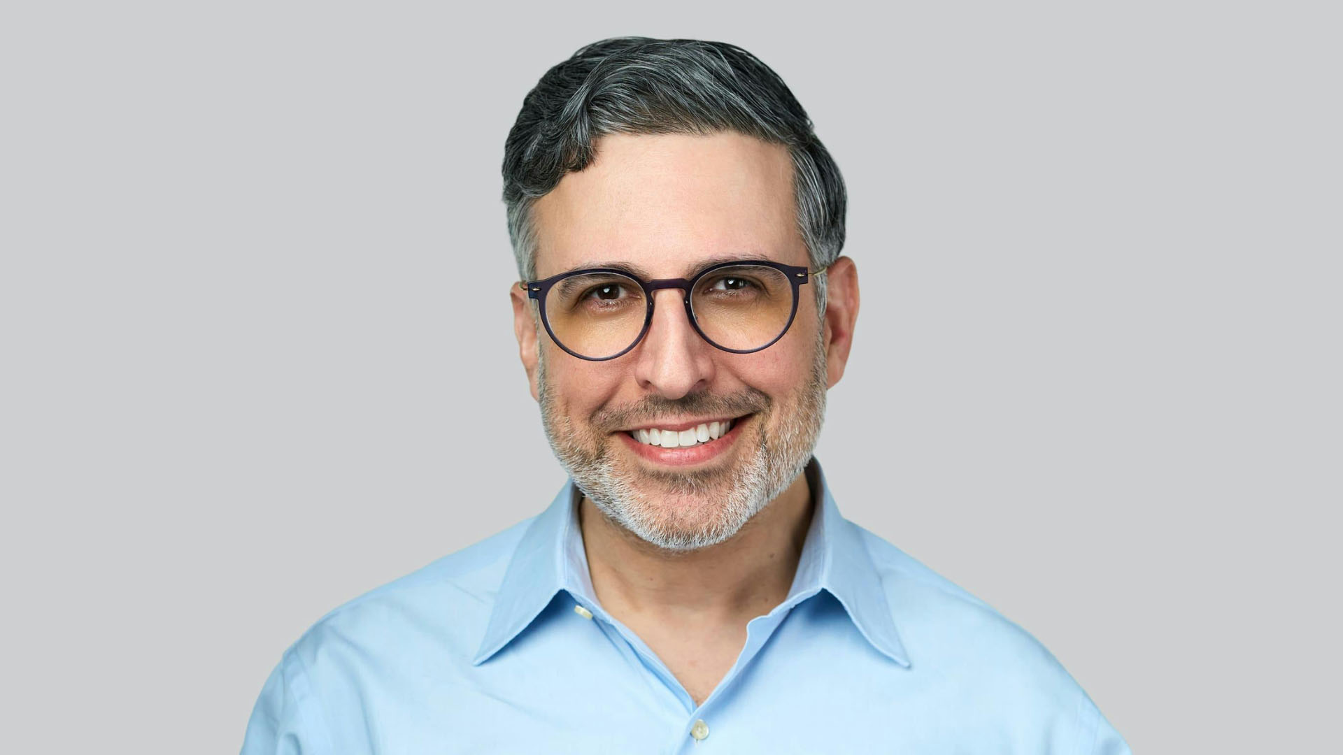 Smiling middle-aged man with grey-streaked hair, round glasses and a light blue shirt against a grey background